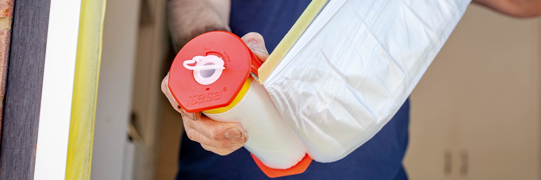 Person holding a roll of masking film with a red handle near a window frame. The window edges are partially covered with yellow tesa tape, presumably for painting or renovations. The person is wearing a dark blue shirt. (This text has been generated by AI)
