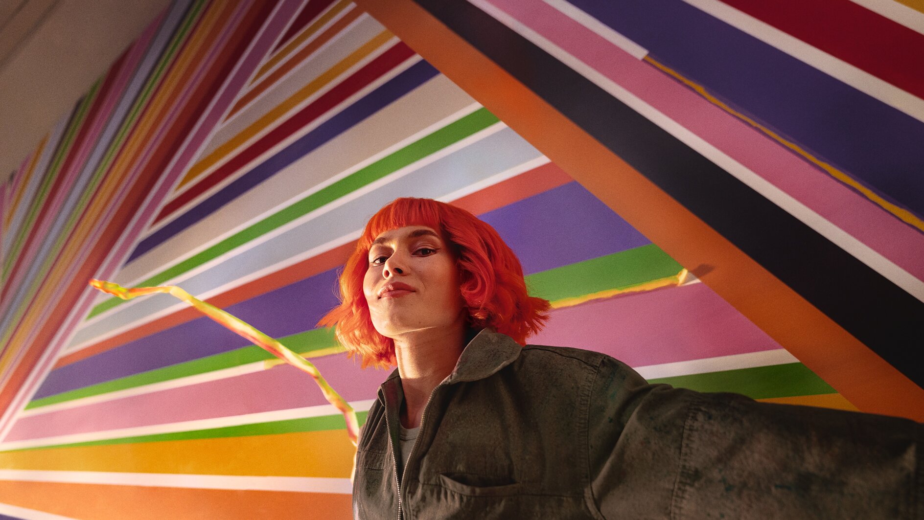 A person with bright red hair stands in front of a colorful geometric mural featuring diagonal stripes in various colors. They are wearing a dark jacket and looking confidently at the camera, with tesa tape prominently used throughout the artwork to create clean and vibrant lines. (This text has been generated by AI)