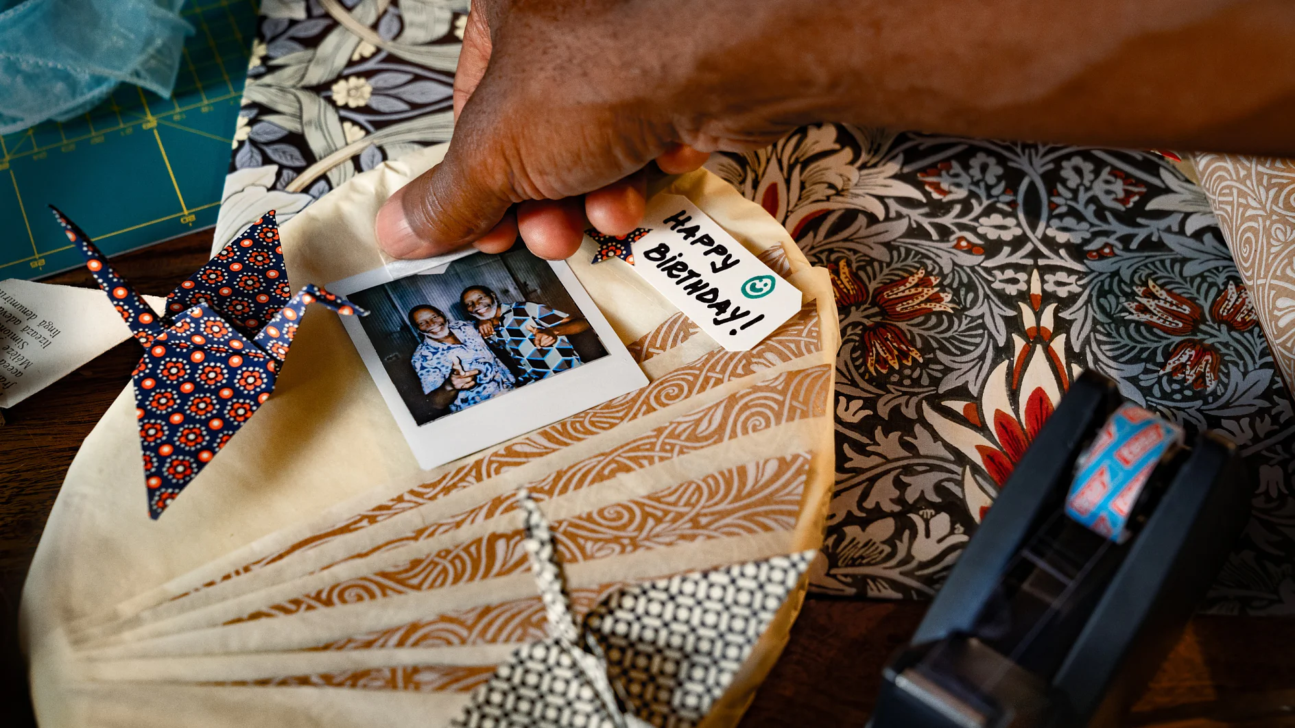 A hand holds a circular fabric piece with a pinned photo of two people beside an origami crane. A tag reads Happy Anniversary. Nearby are patterned papers and a tesa tape dispenser on a wooden surface. (This text has been generated by AI)