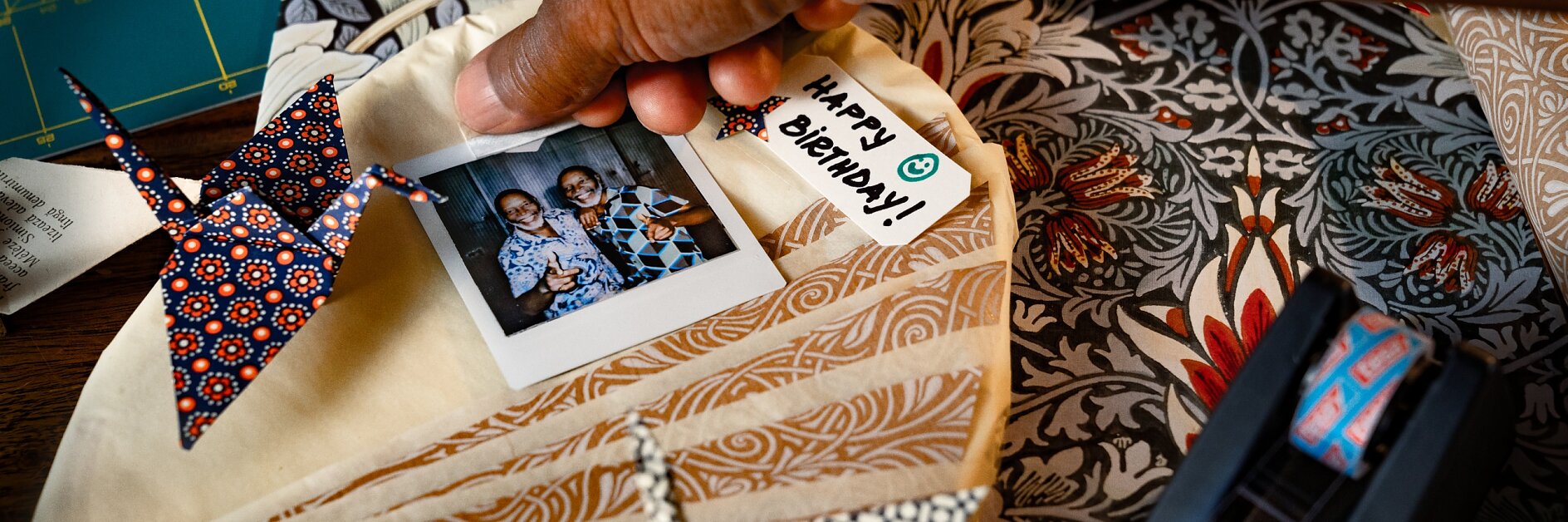 A hand holds a circular fabric piece with a pinned photo of two people beside an origami crane. A tag reads Happy Anniversary. Nearby are patterned papers and a tesa tape dispenser on a wooden surface. (This text has been generated by AI)