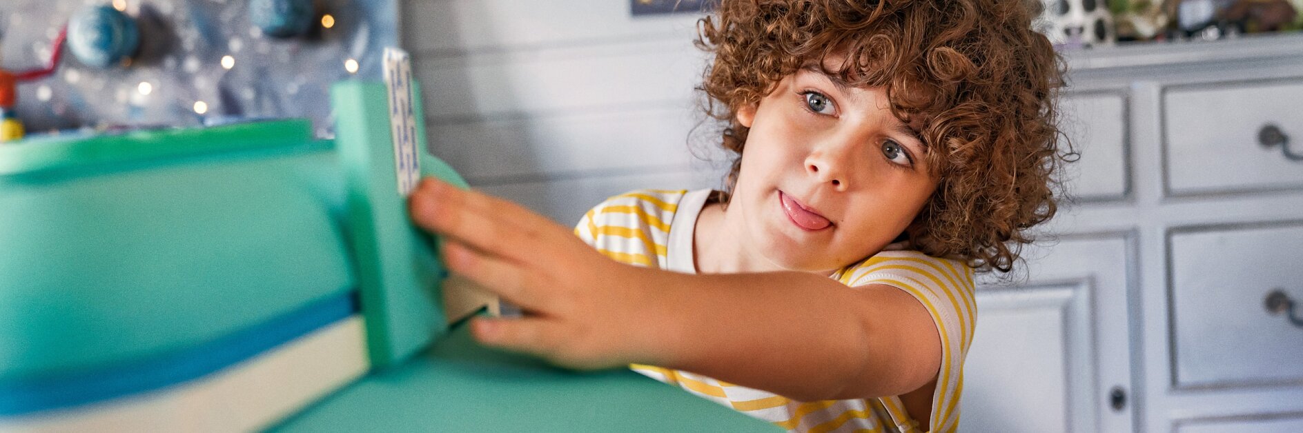 A young child with curly hair and a striped yellow shirt plays with a toy. The room has colorful decorations, a lamp, and a dresser in the background. The child appears focused and has their tongue slightly out. (This text has been generated by AI)