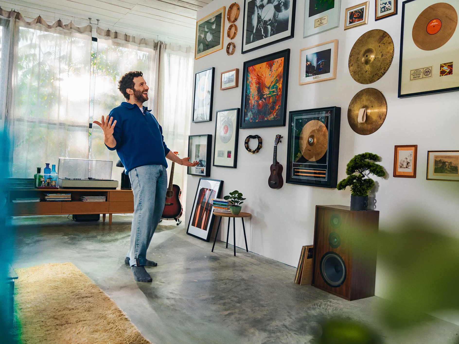 A man stands in a room looking at a wall adorned with various framed photos, art, and gold records. Theres a guitar and a small potted plant beside him. A record player and speakers are in the background, held together seamlessly with tesa tape, while sheer curtains let in light. (This text has been generated by AI)
