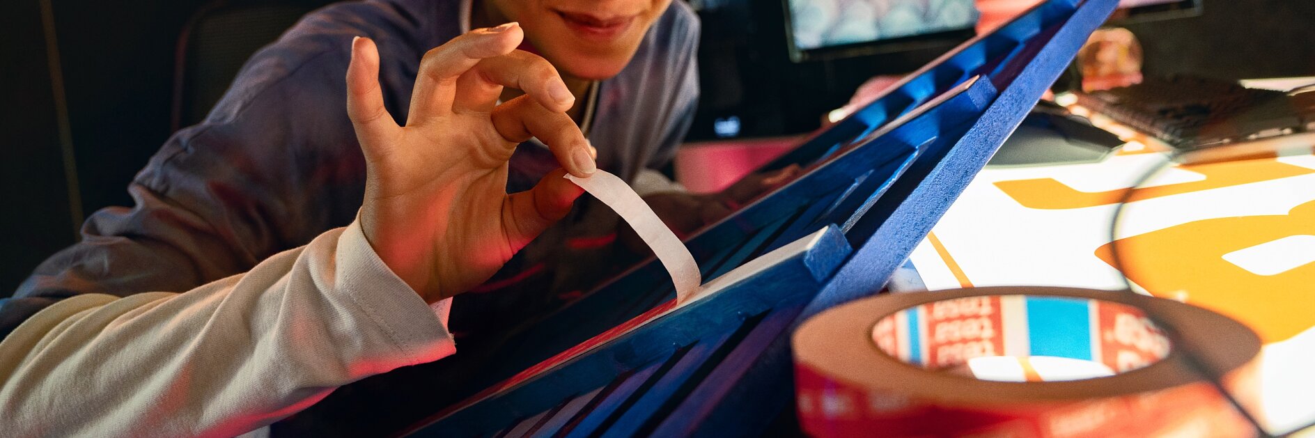 A person with short hair and glasses is seated at a desk, peeling tesa tape from a blue object. In the background, a computer monitor displays colorful graphics. Rolls of tesa tape and papers are visible on the desk. (This text has been generated by AI)