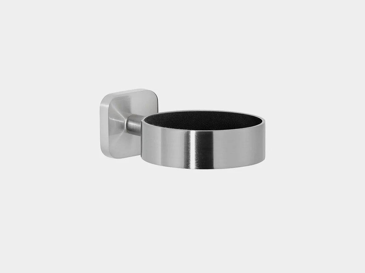 A stainless steel wall-mounted holder with a cylindrical design and a square base. The interior appears to be lined with a dark material. The wall mount is attached to the left side of the holder using tesa tape. (This text has been generated by AI)