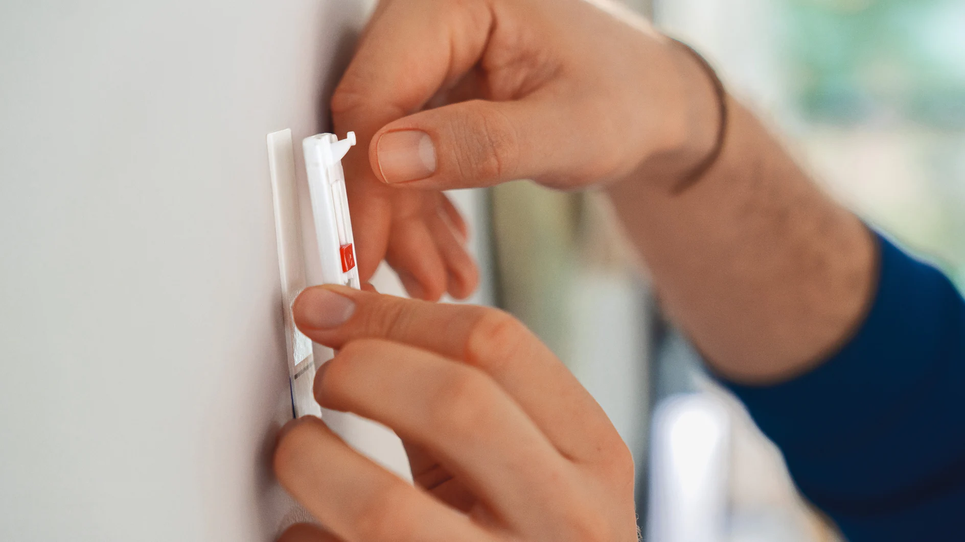 A person is adjusting or installing a white wall-mounted device with a red button using tesa tape. The hands are shown in close-up, and the wall is light-colored. The person is wearing a blue sleeve. (This text has been generated by AI)