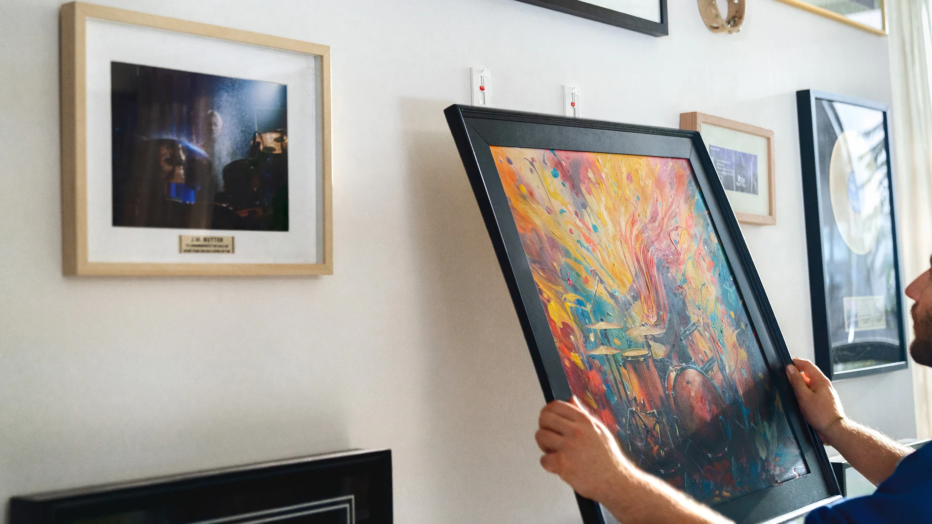 A person is hanging a colorful abstract painting on a wall. The wall already has several framed pictures, including one featuring a dark, ambiguous scene. The persons arm and part of their torso are visible on the right side of the image, and they are using tesa tape to affix the new painting. (This text has been generated by AI)