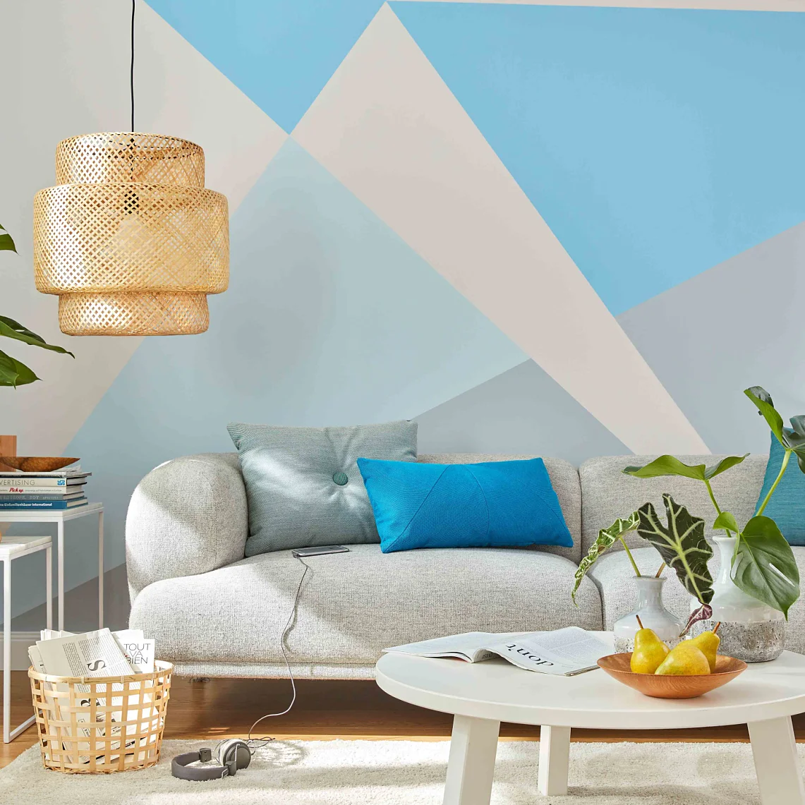 Ein modernes Wohnzimmer mit einem hellgrauen Sofa, blauen und blaugrünen Kissen und einer geometrischen blau-weißen Wand. Darüber hängt eine Hängelampe aus Korbgeflecht. Auf einem beigen Teppich steht ein runder weißer Couchtisch mit Pflanzen und Obst. Auf einem weißen Beistelltisch steht eine Topfpflanze. (This text has been generated by AI)