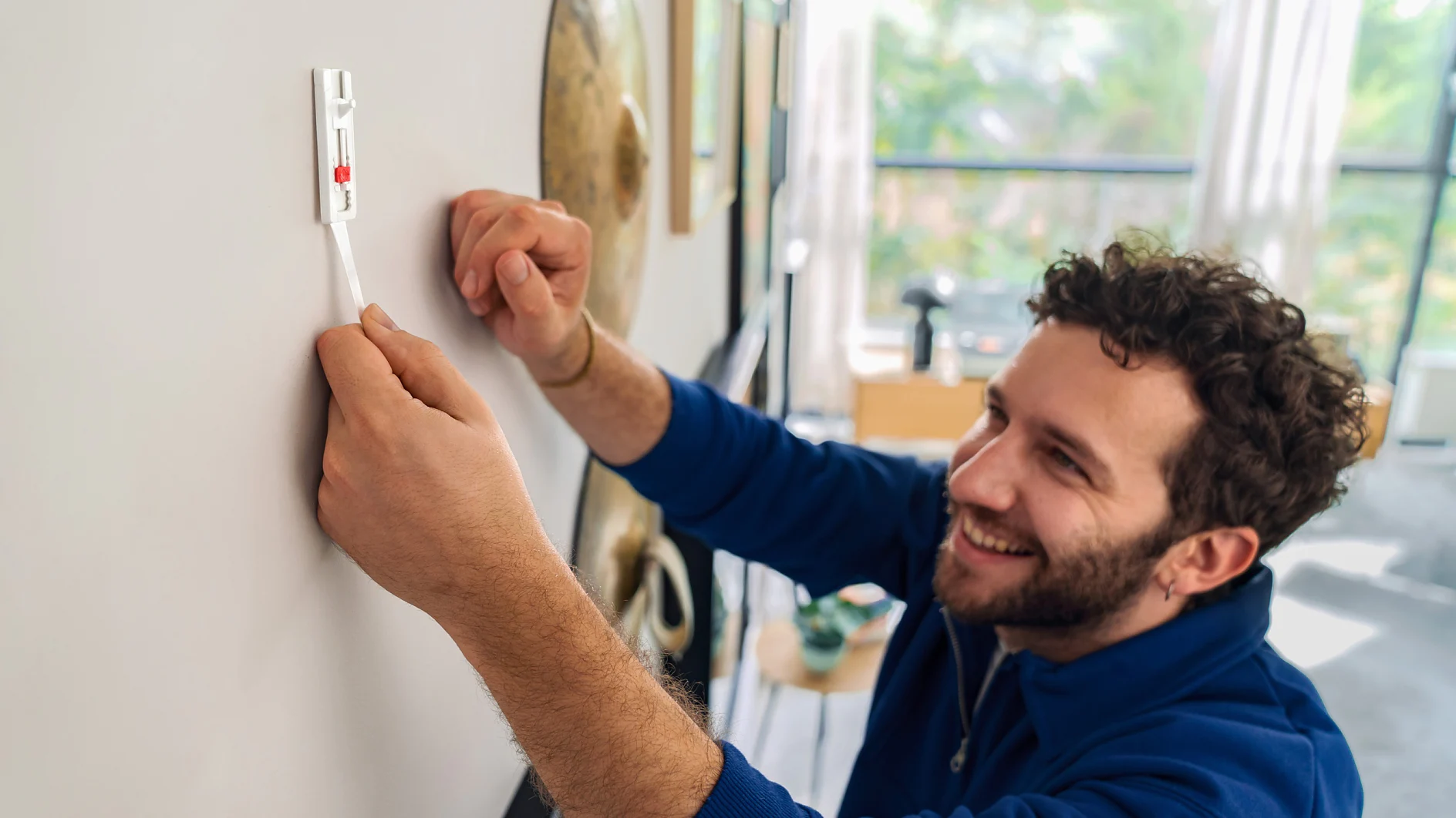 A man with curly hair and a beard is smiling while using a level tool to hang something on a white wall in a home setting. He is using tesa tape to assist with the task. He is wearing a blue shirt, and there are blurred images of furniture and decor in the background. (This text has been generated by AI)