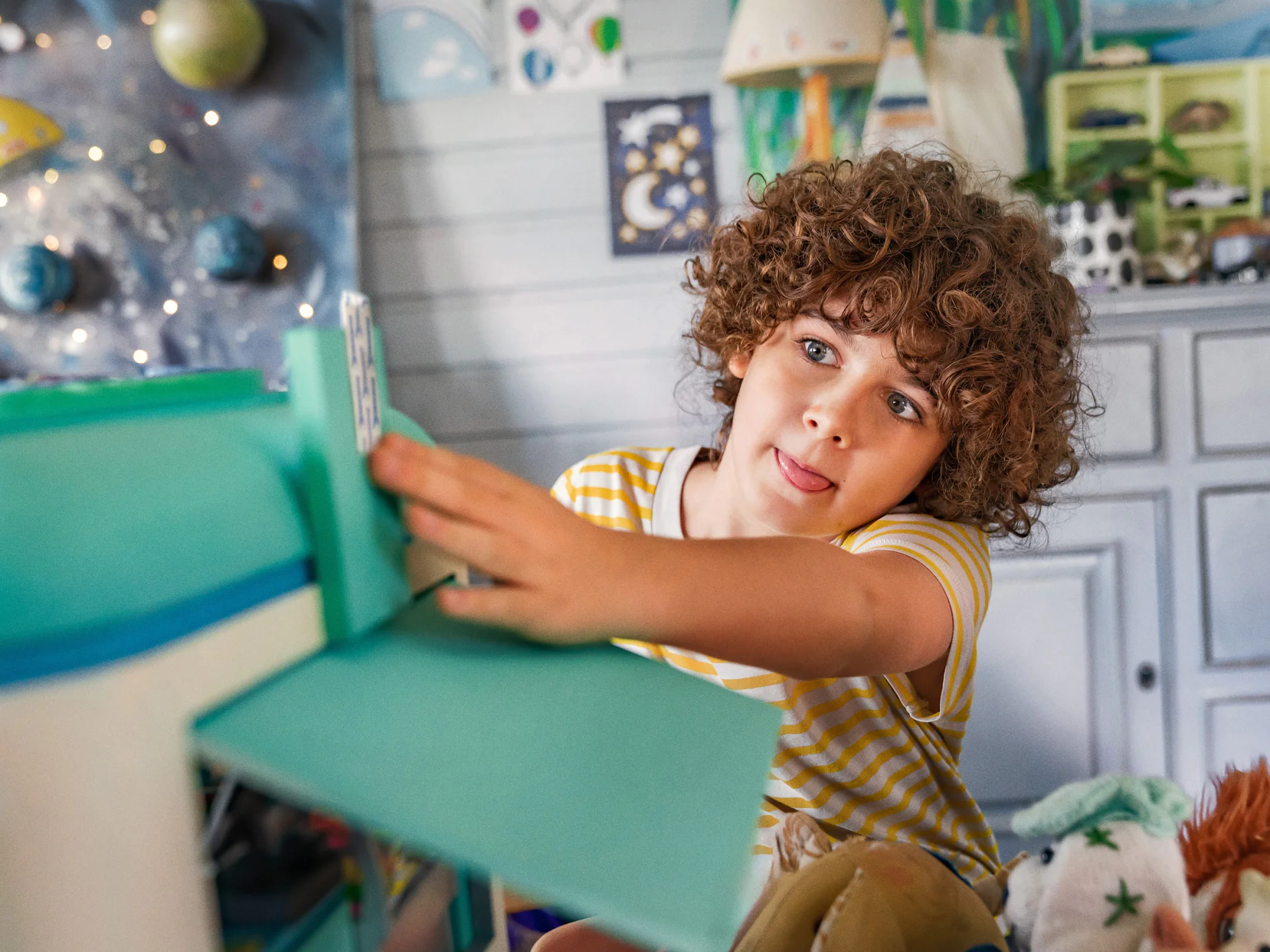 A child with curly hair, wearing a yellow striped shirt, is playing with a toy house. They have an attentive expression and are surrounded by other toys. The room has a playful decor, including stars and planets on the wall. (This text has been generated by AI)