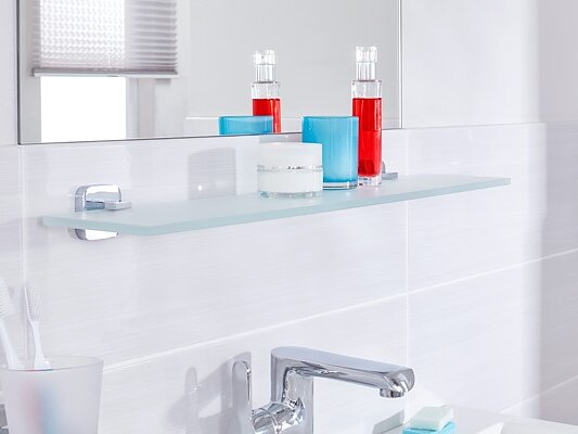 A modern bathroom showcases a glass shelf with blue and white candles alongside two red bottles. Below the shelf, a sink with a chrome faucet is partially visible. Toothbrushes in a holder are positioned on the left, while a hairdryer is mounted on the right using tesa tape. (This text has been generated by AI)