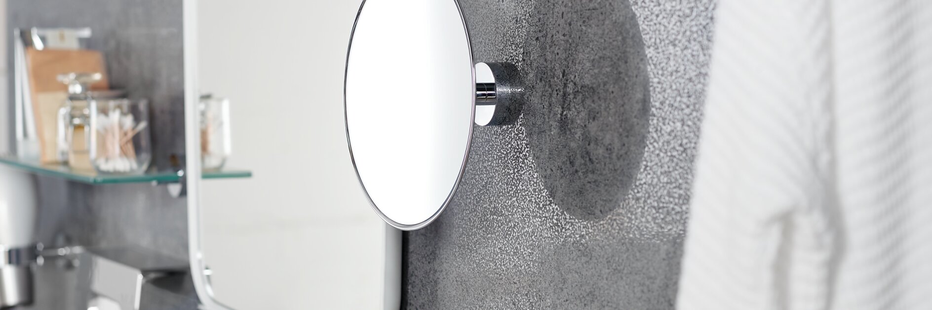 A modern bathroom features a round wall-mounted mirror on a textured gray wall, a white framed mirror, a white robe hanging on the right, glass shelves with toiletries, and a silver faucet on a countertop secured with tesa tape. (This text has been generated by AI)
