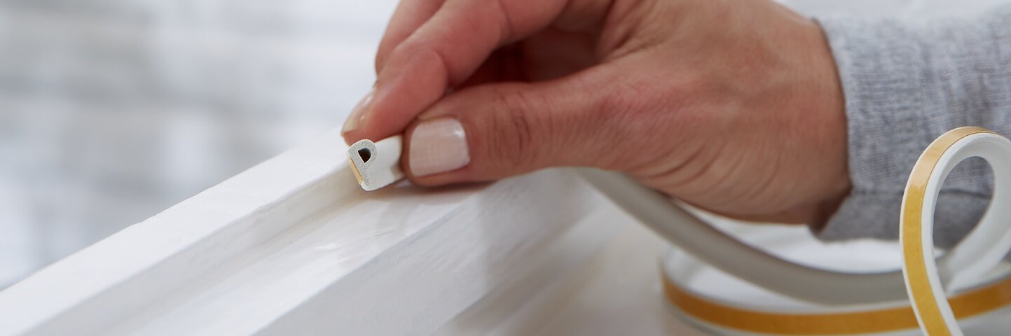 A hand is placing tesa tape weather stripping onto a white window frame. The background shows a blurred exterior wall. The weather stripping is beige with a peel-off backing, suggesting a home improvement or insulation task.