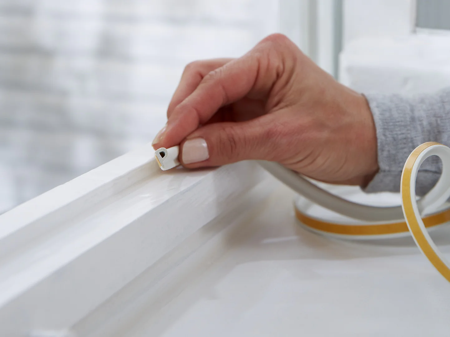 A hand is placing tesa tape weather stripping onto a white window frame. The background shows a blurred exterior wall. The weather stripping is beige with a peel-off backing, suggesting a home improvement or insulation task. (This text has been generated by AI)