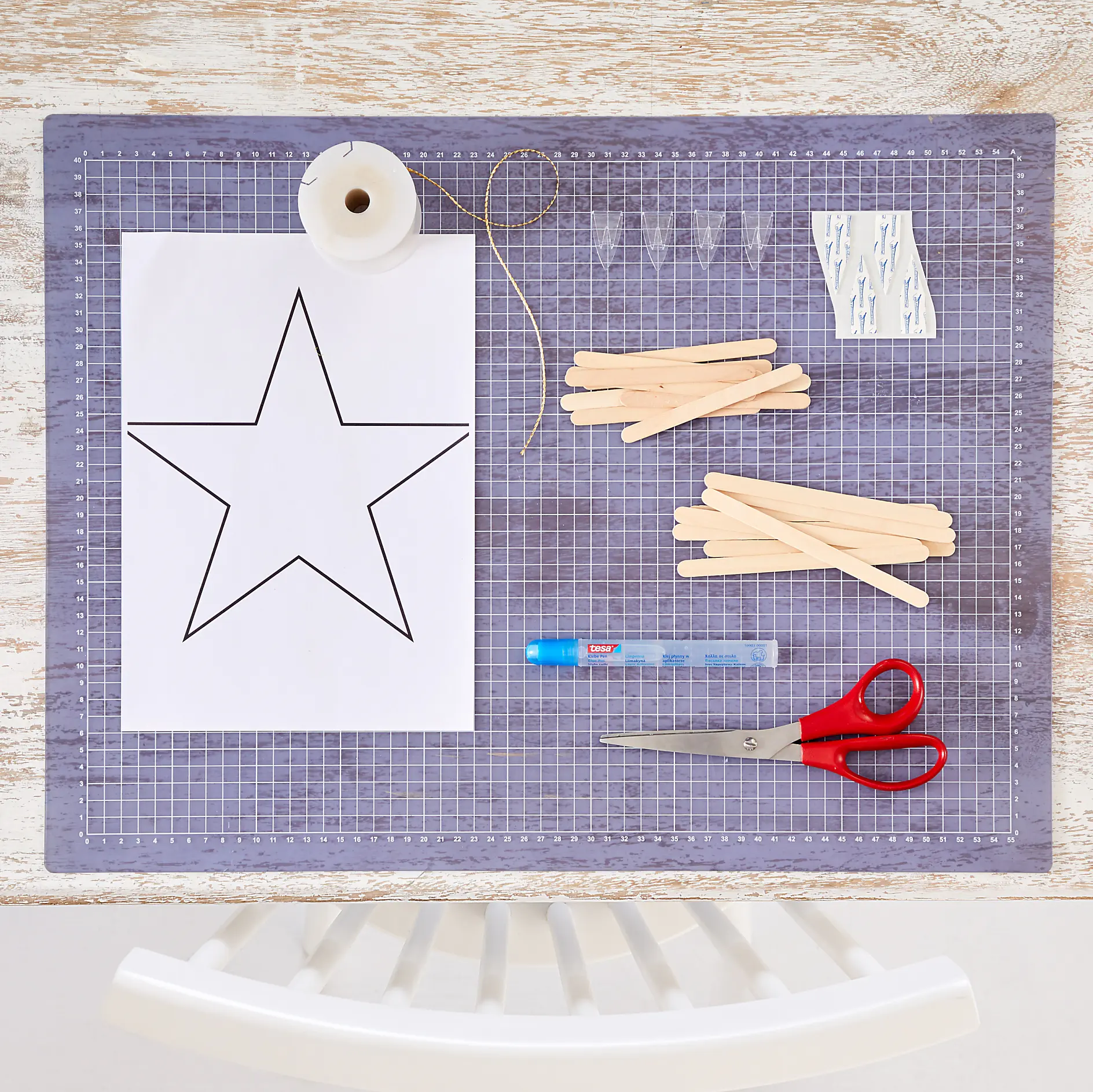 A crafting setup on a table includes a printed star outline on paper, a spool of thread, wooden sticks, a glue stick, scissors, tesa tape adhesive hooks. Everything is arranged neatly on a cutting mat. A white chair is partially visible at the bottom. (This text has been generated by AI)