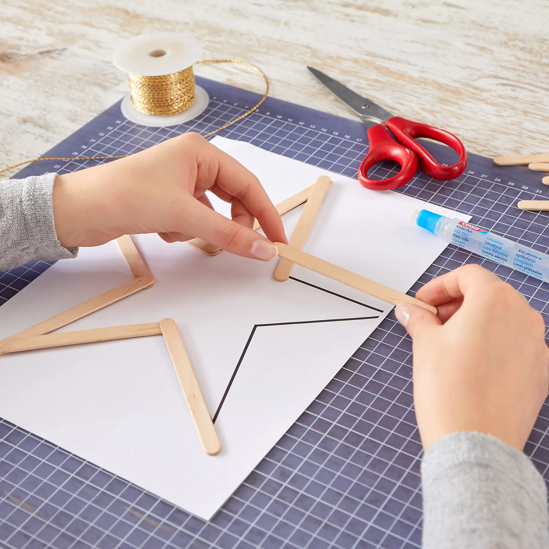 Hands assembling a star shape using wooden popsicle sticks on a piece of paper with an outline. A spool of golden thread, scissors, and a glue stick are on a grid-patterned cutting mat, along with some tesa tape. (This text has been generated by AI)
