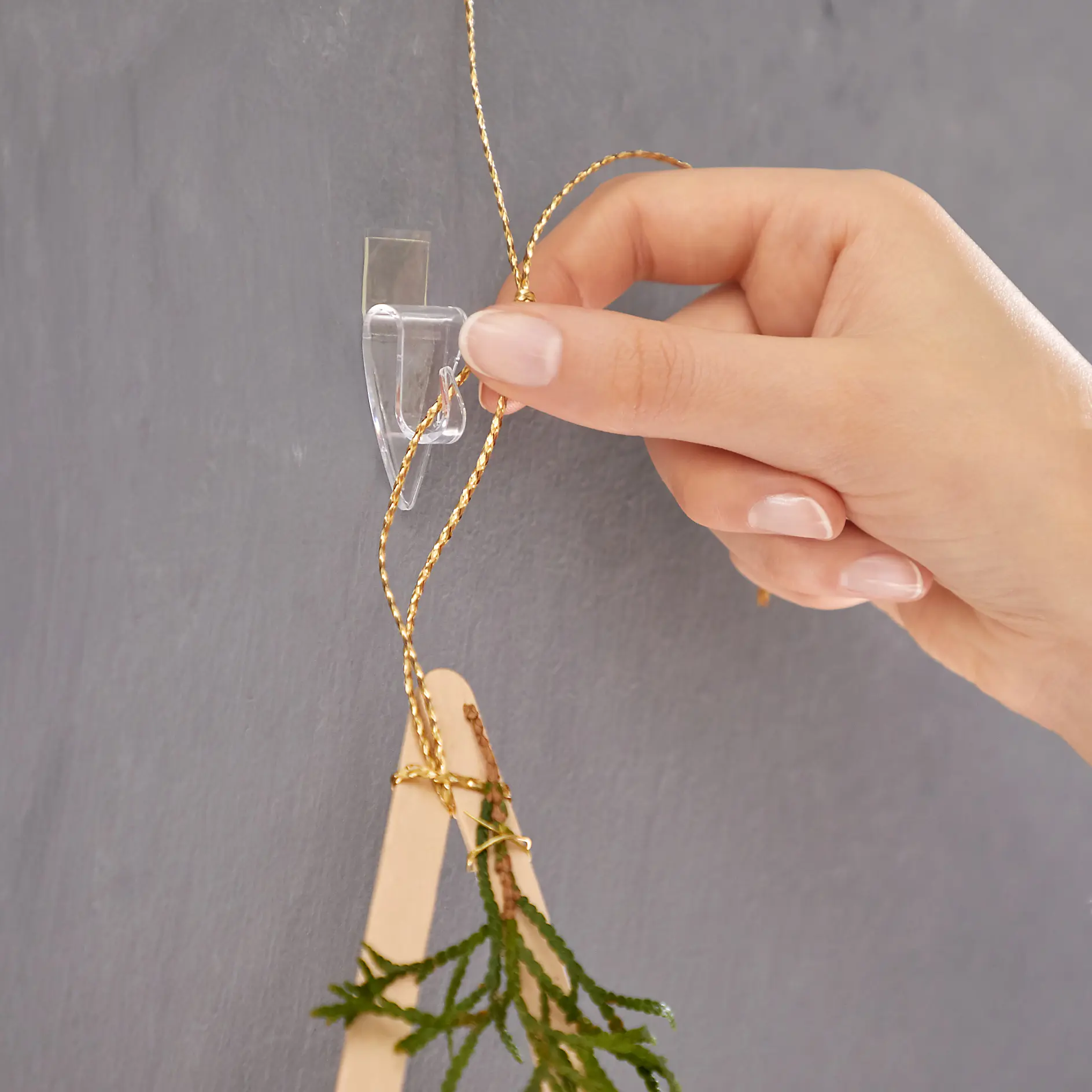 A hand is attaching a string of gold-colored beads and a small wooden ornament with greenery to a clear adhesive wall hook using tesa tape. The background is a smooth, gray wall surface. (This text has been generated by AI)