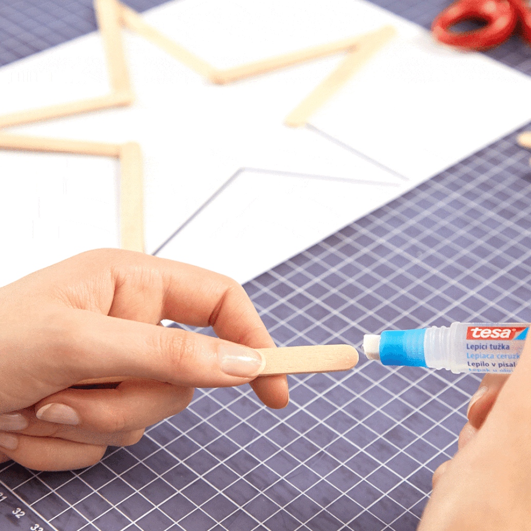 A person applying glue from a tube onto the end of a wooden craft stick, with other sticks arranged in a star shape on a piece of paper in the background. The scene is on a cutting mat with a grid pattern alongside tesa tape. (This text has been generated by AI)