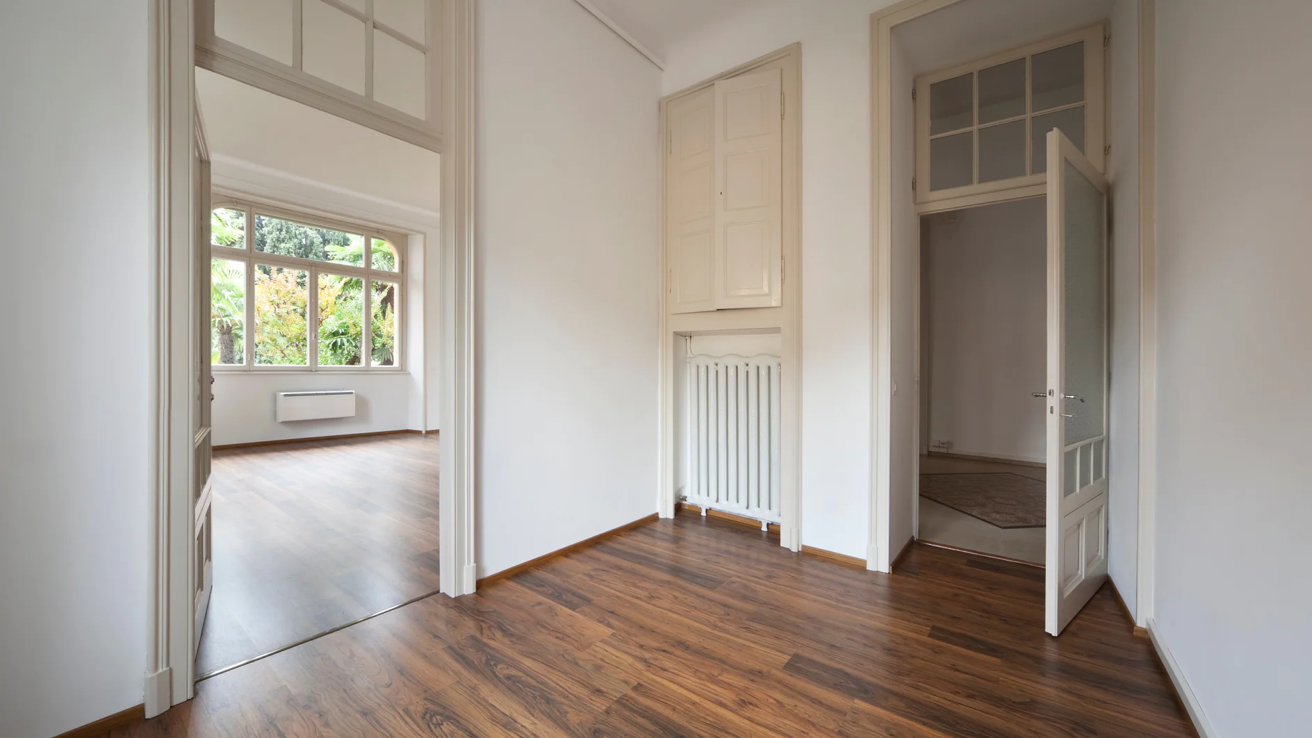 Ein leerer Raum mit Holzboden und weißen Wänden verfügt über eine Tür, die zu einem anderen Raum mit einem großen Fenster führt. Auf der rechten Seite befindet sich ein Heizkörper und eine offene Tür mit Oberlichtfenstern darüber. Der Raum ist von natürlichem Licht durchflutet. Tesa-Klebeband kann nützlich sein, um Gegenstände in diesem hellen, offenen Bereich zu befestigen, ohne Spuren auf den glatten Oberflächen zu hinterlassen. (This text has been generated by AI)