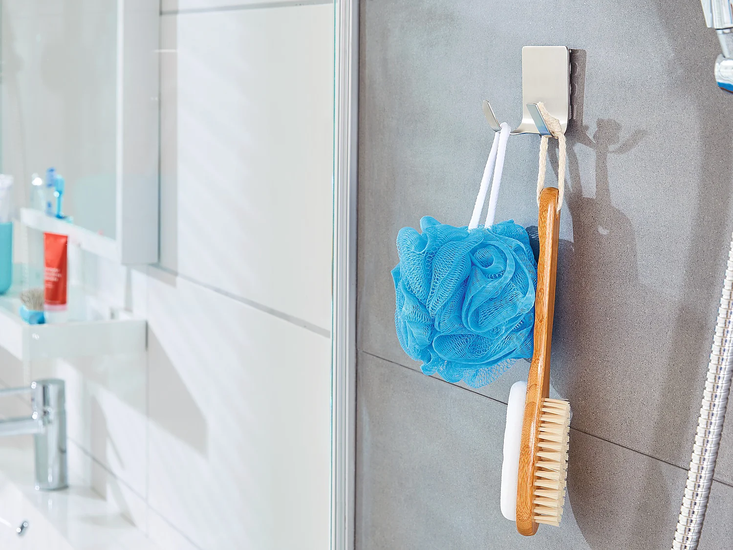 Ein Duschbereich im Badezimmer mit grau gefliester Wand. Ein blauer Duschschwamm und eine Holzbürste hängen an Haken, die mit Tesa-Klebeband befestigt sind. Rechts ist ein Duschkopf zu sehen. Im Hintergrund steht auf einer Ablage ein weißes Waschbecken mit verschiedenen Toilettenartikeln. (This text has been generated by AI)