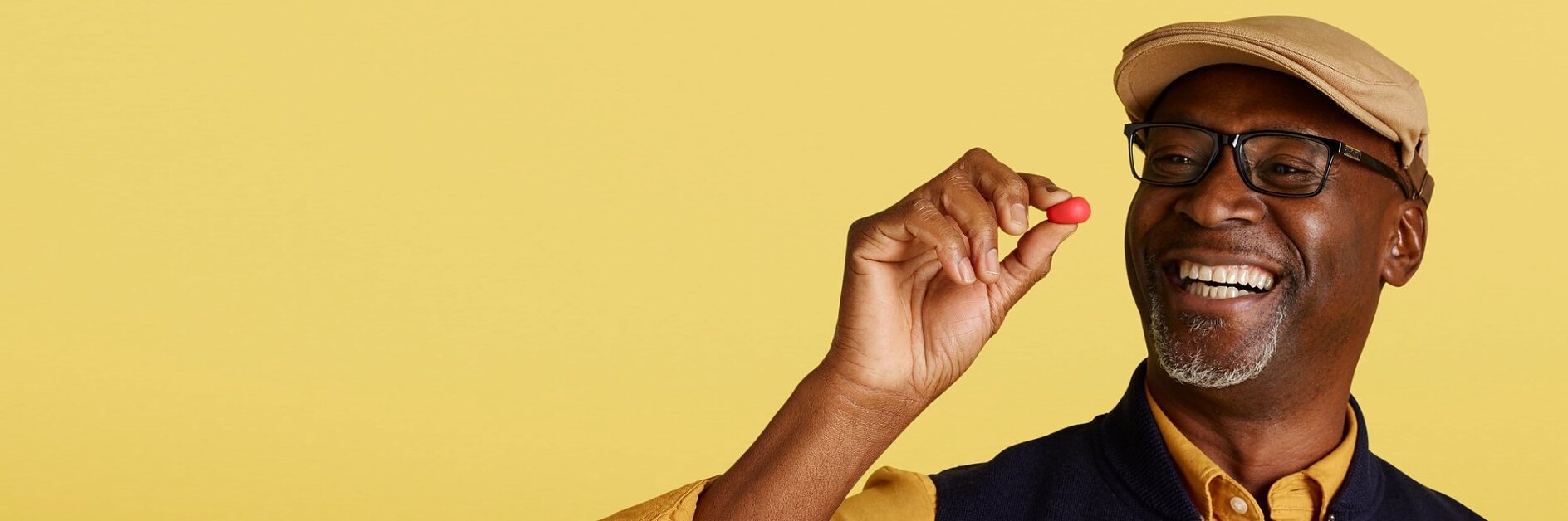 A man wearing a cap, glasses, and a casual shirt with a vest is smiling while holding a small red object between his fingers. The background is a solid light yellow color. There are no references to tape in the provided text that would require replacing with tesa tape. (This text has been generated by AI)