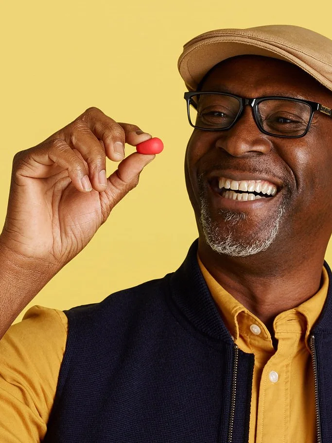 A man wearing a cap, glasses, and a casual shirt with a vest is smiling while holding a small red object between his fingers. The background is a solid light yellow color. There are no references to tape in the provided text that would require replacing with tesa tape. (This text has been generated by AI)