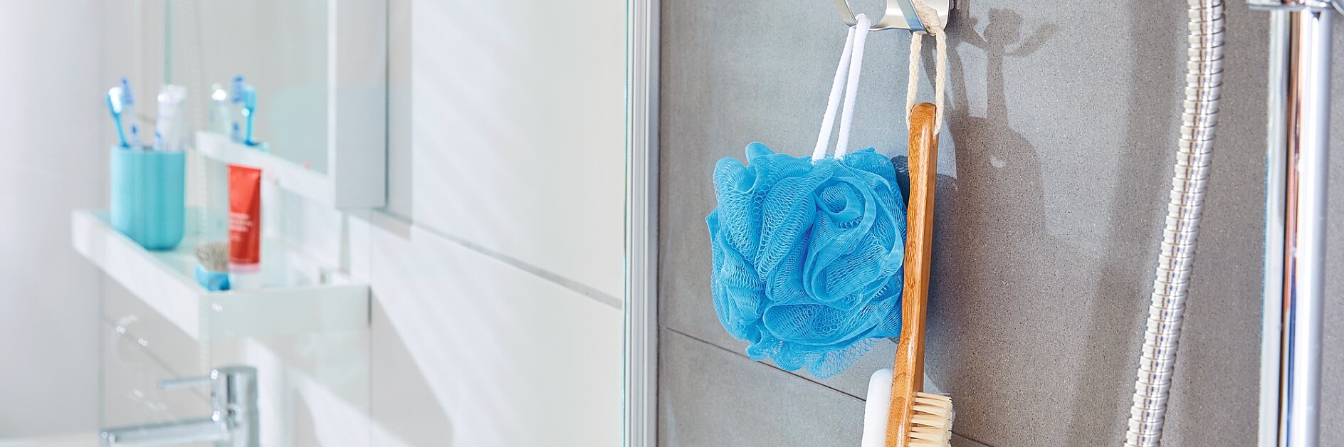 Eine Badezimmerszene zeigt eine Wand, an der ein blauer Luffaschwamm und eine hölzerne Badebürste an Haken hängen, die mit Tesa-Klebeband befestigt sind. Rechts ist ein Duschkopf zu sehen. Im Hintergrund steht ein Waschbecken mit Toilettenartikeln auf einer weißen Arbeitsplatte. (This text has been generated by AI)