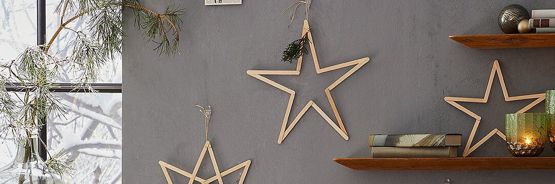Sternförmige Holzdekorationen hängen an einer grauen Wand, mit Tesa-Klebeband befestigt und mit kleinen Tannenzweigen geschmückt. Auf Holzregalen stehen gestapelte Bücher und ein grüner Kerzenständer. Ein Fenster gibt den Blick auf eine verschneite Landschaft draußen frei. (This text has been generated by AI)