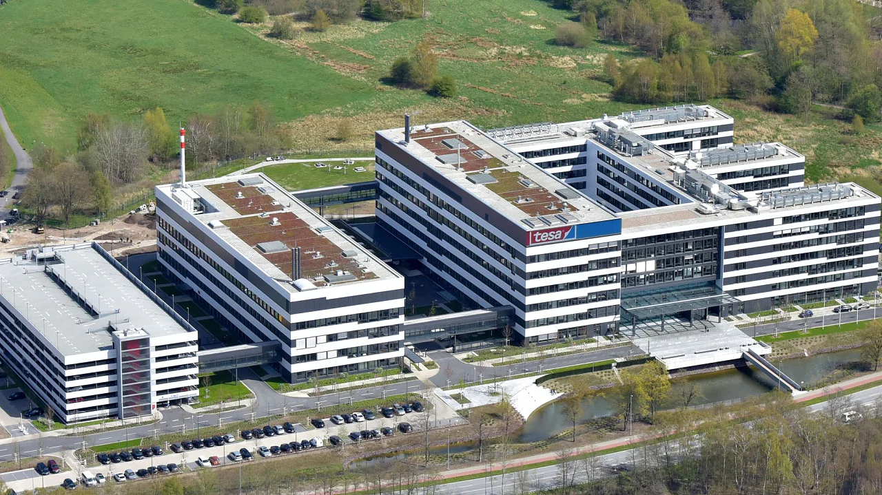 Aerial view of a large, modern office complex surrounded by green spaces and a pond. Residential houses are visible in the background, alongside roads and construction areas. Trees surround the area, creating a mix of urban and natural elements. (This text has been generated by AI)