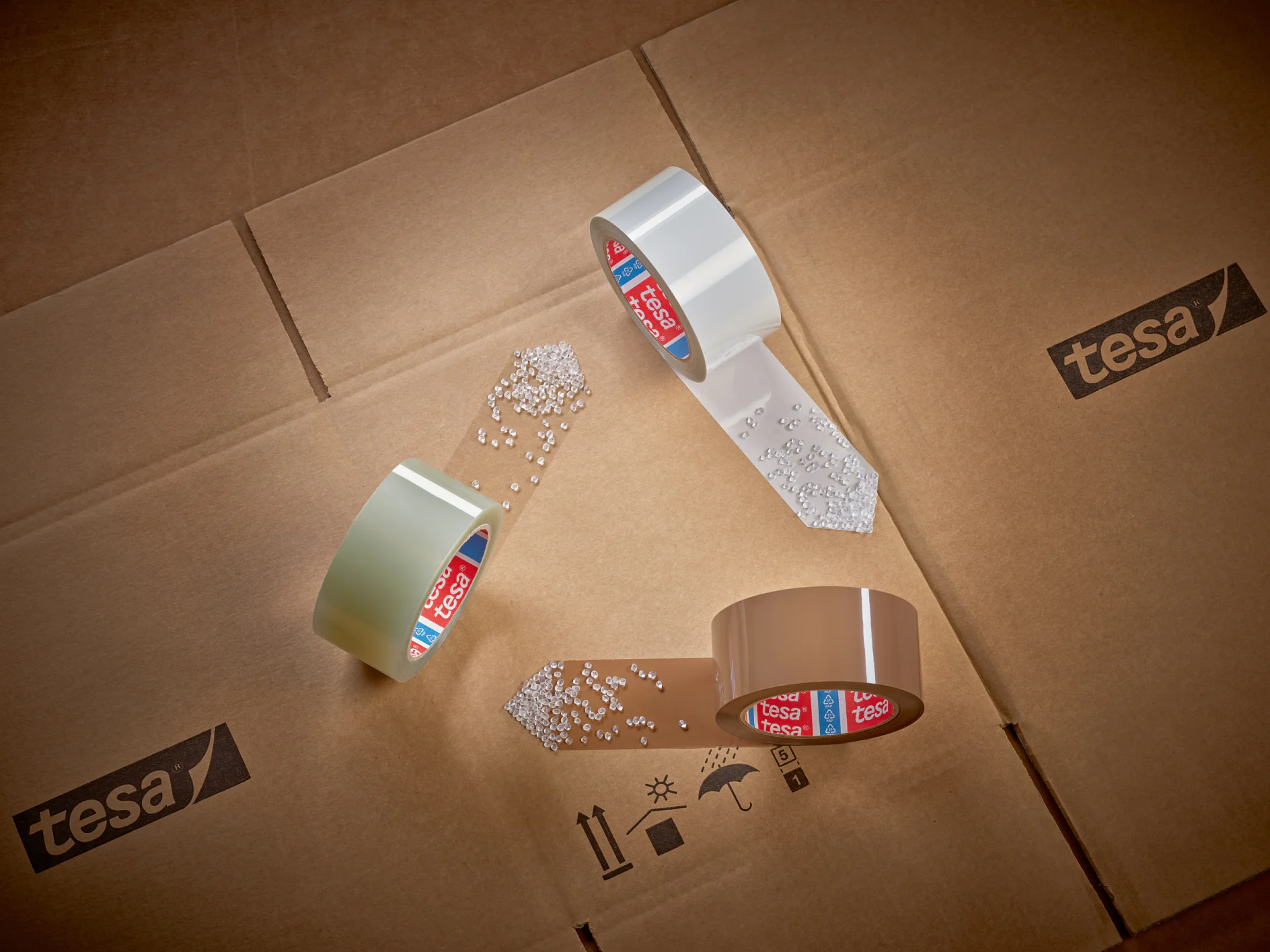 tesa-ID-60412-recycled-PET-packaging-tape-family-mood%20%282%29-Print Three rolls of tesa tape are arranged in a triangular formation on cardboard boxes. White pellets are scattered near the ends of the tesa tape. Tesa branding is visible on the tesa tape cores and the boxes. (This text has been generated by AI)