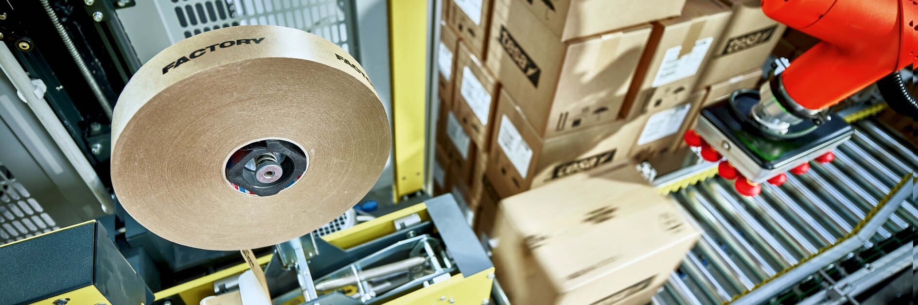 In a factory setting, theres a roll of tesa tape labeled Factory near a packaging machine. A red robotic arm maneuvers a brown cardboard box on a conveyor belt. Stacked boxes marked with tesa are visible in the background.