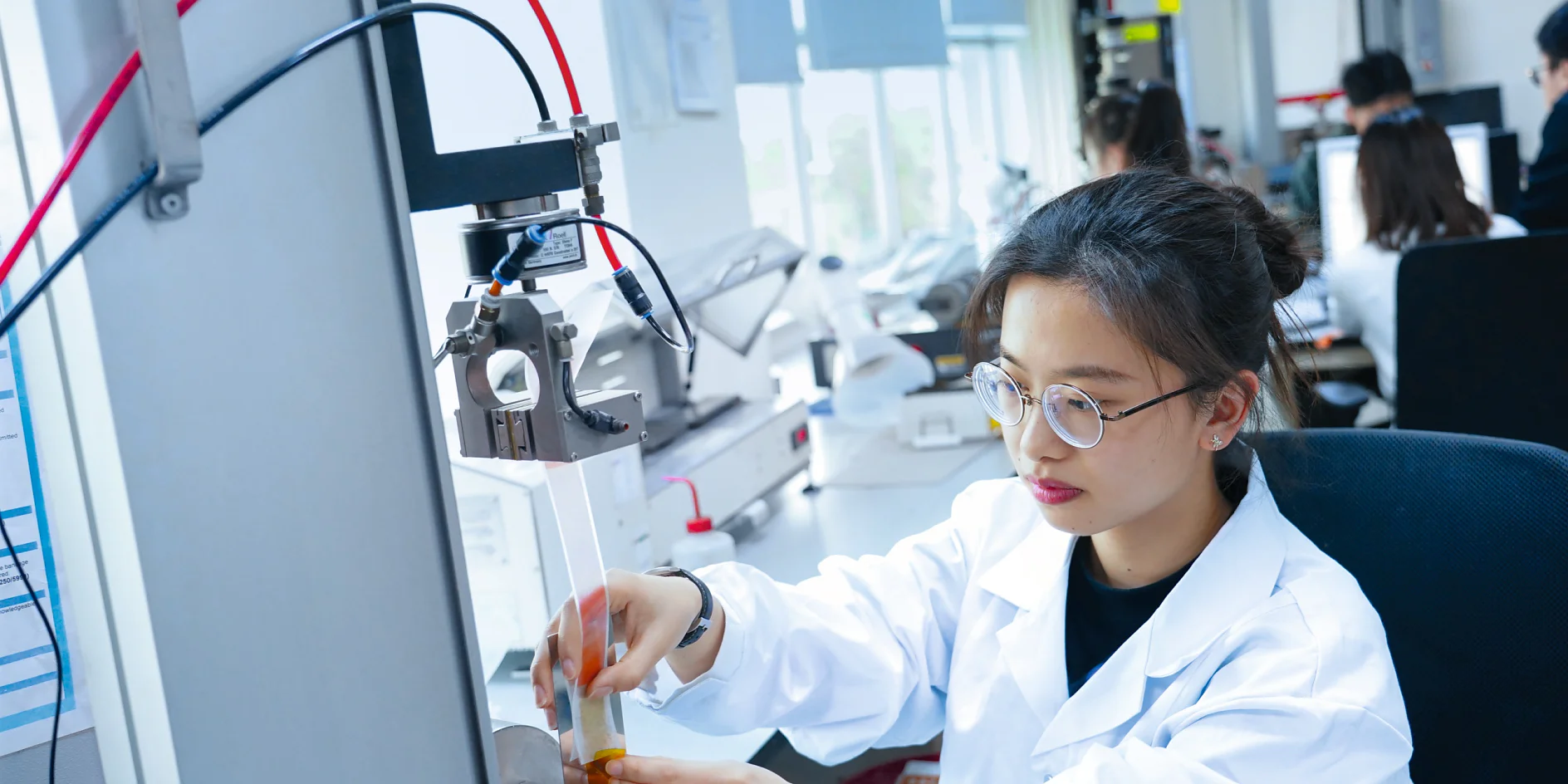 A person in a lab coat and glasses operates equipment in a laboratory. They are adjusting a component near a machine with wires using tesa tape. Other people are working in the background, and computer monitors are visible. The setting is brightly lit and organized. (This text has been generated by AI)