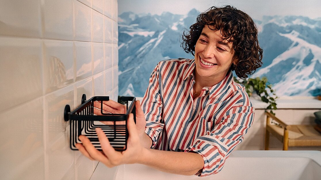 A person with curly hair in a striped shirt mounts a black wire rack onto a white tiled wall using tesa tape. A painted mountainous landscape is visible in the background. (This text has been generated by AI)