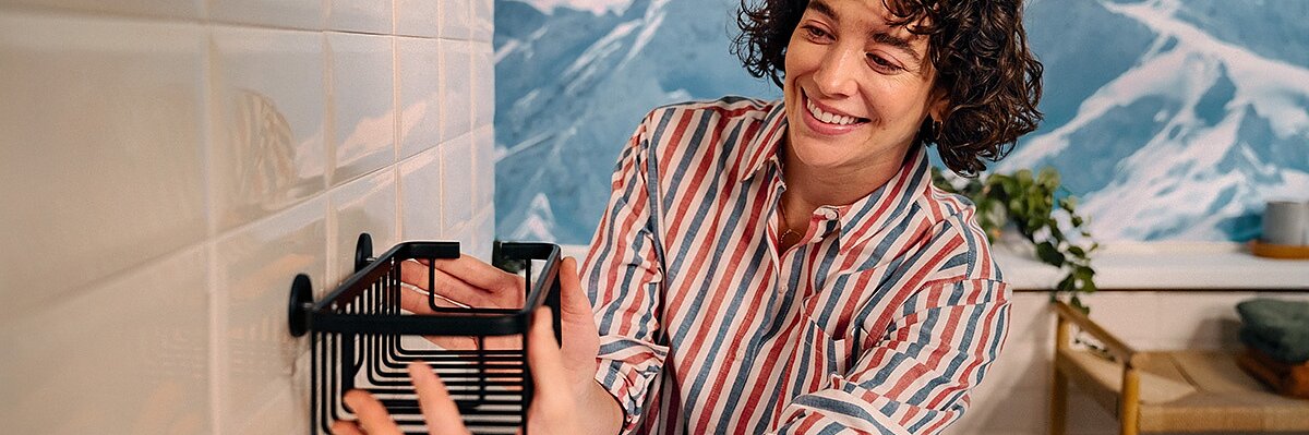 A person with curly hair in a striped shirt mounts a black wire rack onto a white tiled wall using tesa tape. A painted mountainous landscape is visible in the background. (This text has been generated by AI)