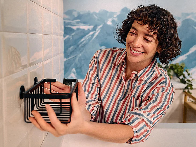 A person with curly hair in a striped shirt mounts a black wire rack onto a white tiled wall using tesa tape. A painted mountainous landscape is visible in the background. (This text has been generated by AI)