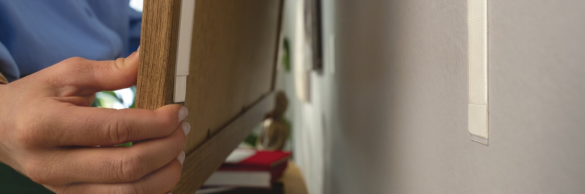 A person is mounting a wooden frame on a white wall using tesa tape strips. They are pressing the frame against the wall. There are books and a small decorative item on a wooden surface below the frame.