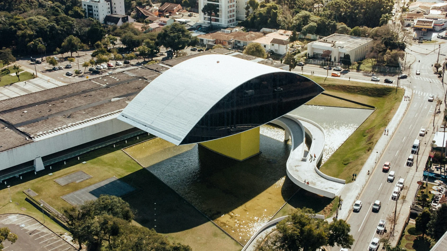 Oscar Niemeyer Museum 3 Aerial view of the Oscar Niemeyer Museum in Curitiba, Brazil. The building features a distinctive eye-shaped structure with a reflective facade, surrounded by a green lawn. Roads with traffic and surrounding residential areas are visible. (This text has been generated by AI)