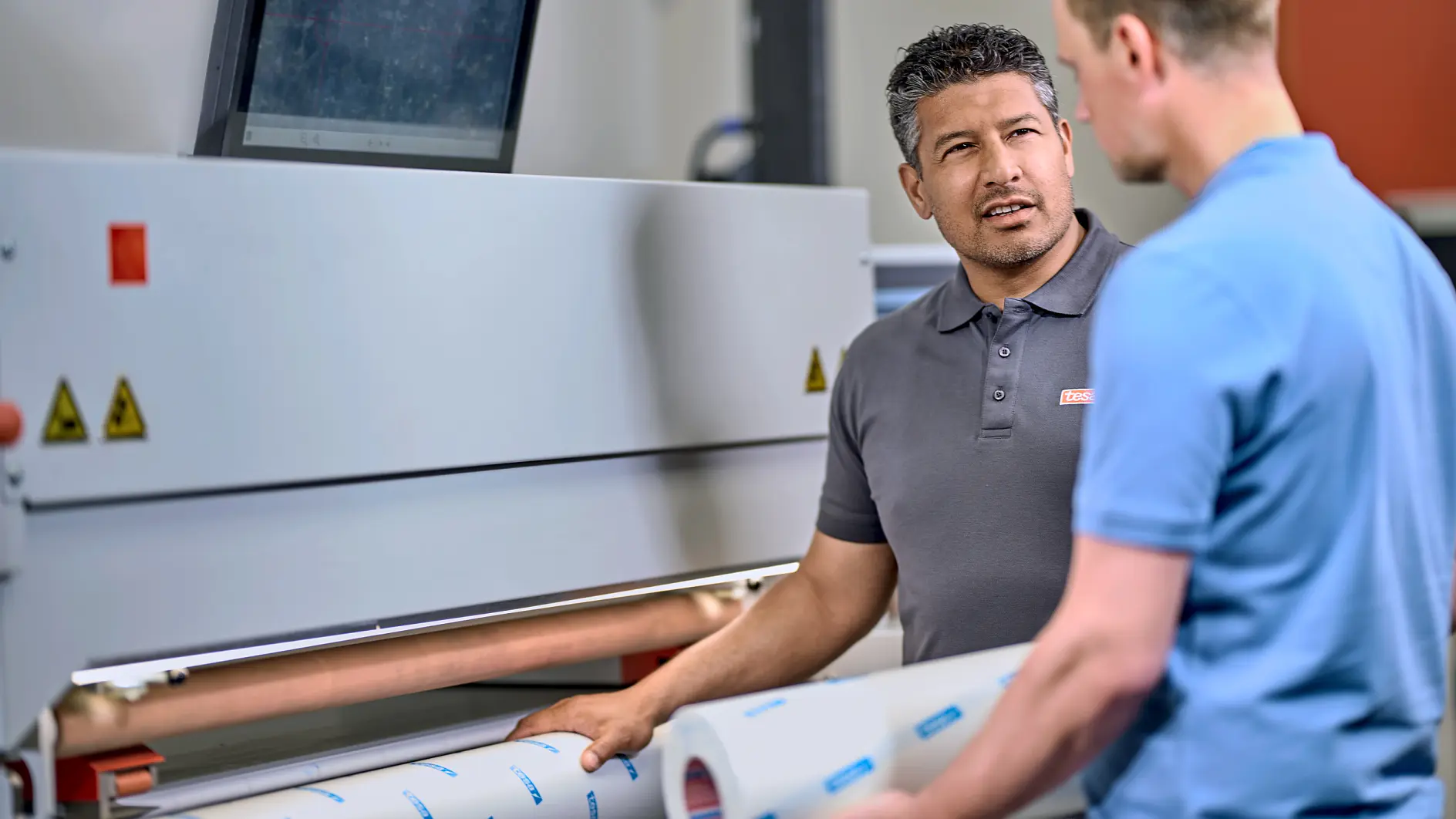 Two men are standing in front of a large industrial machine, discussing something. One man is holding a roll of tesa tape. A computer monitor is mounted on top of the machine. They are in a well-lit workspace.