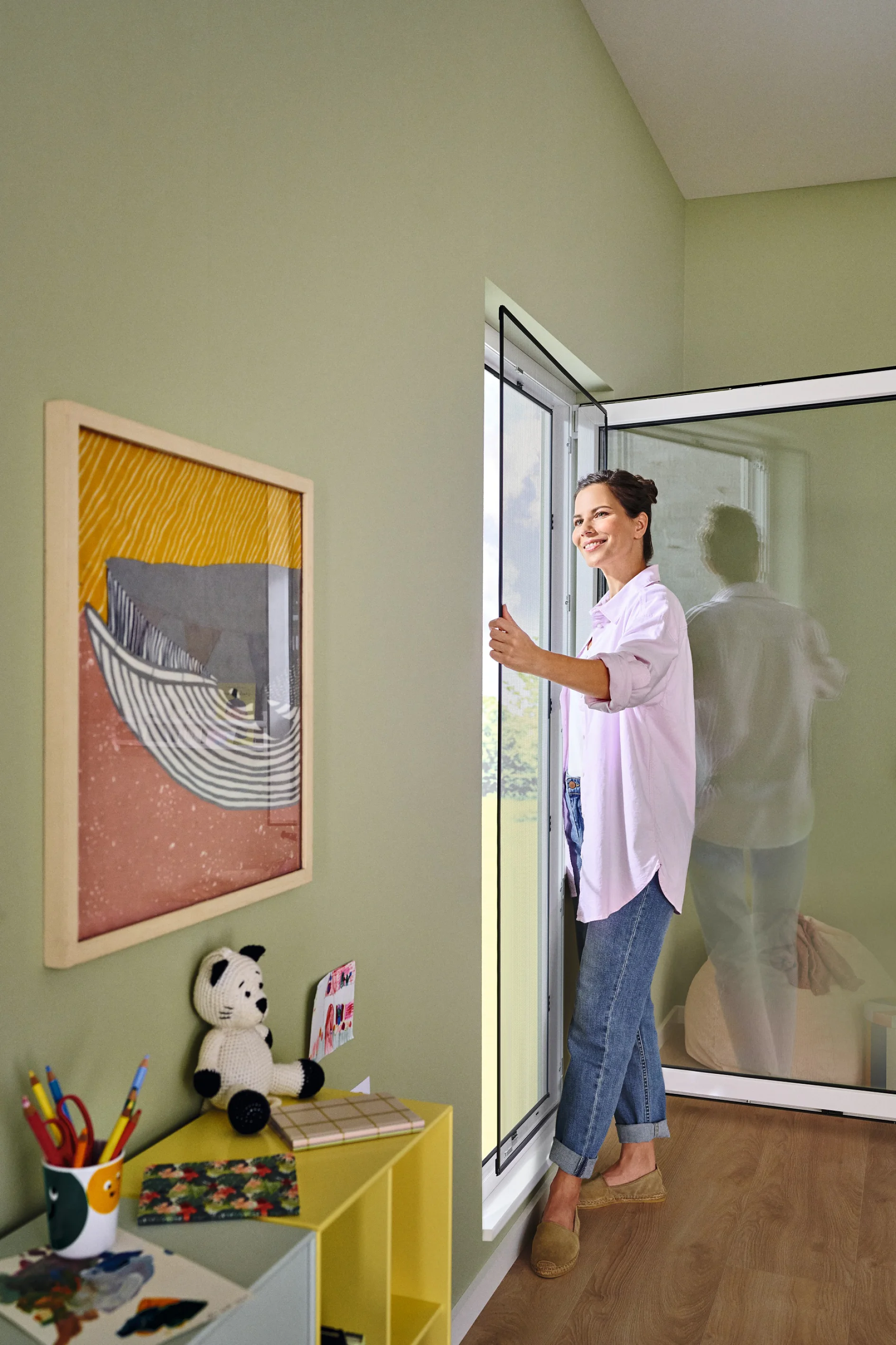 A woman in a white shirt and jeans stands by a large sliding glass door, letting in light. A framed abstract artwork hangs on the pale green wall. A yellow shelf with a stuffed toy, books, and colored pencils secured using tesa tape is in the foreground. (This text has been generated by AI)