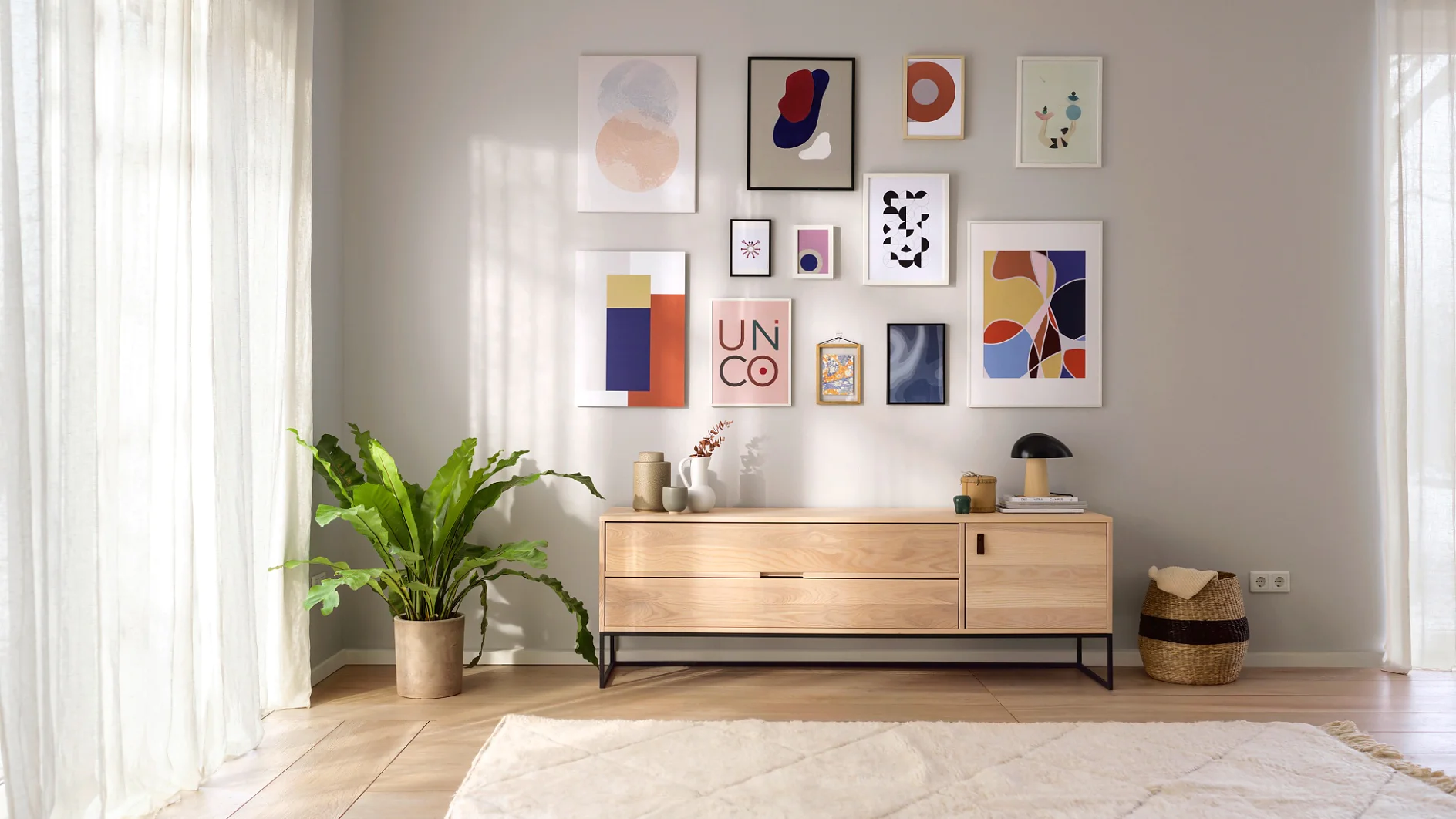 Ein modernes Wohnzimmer mit hellen Wänden, einem großen Teppich und einem Sideboard aus Holz. Abstrakte Kunstdrucke sind mit Tesa-Klebeband an der Wand über dem Sideboard befestigt. Links steht eine Topfpflanze, rechts ein Korb. Sonnenlicht durchflutet den Raum. (This text has been generated by AI)