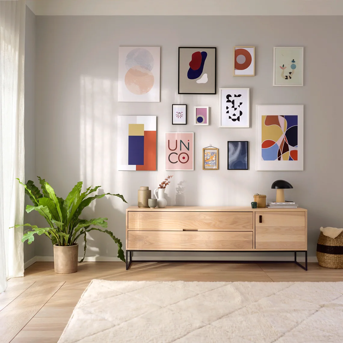 Ein modernes Wohnzimmer mit hellen Wänden, einem großen Teppich und einem Sideboard aus Holz. Abstrakte Kunstdrucke sind mit Tesa-Klebeband an der Wand über dem Sideboard befestigt. Links steht eine Topfpflanze, rechts ein Korb. Sonnenlicht durchflutet den Raum. (This text has been generated by AI)
