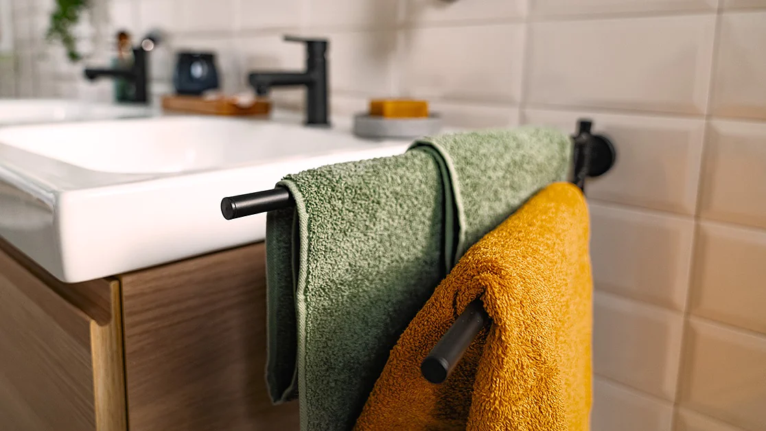Zwei Handtücher, ein grünes und ein orangefarbenes, hängen an einem schwarzen Handtuchhalter, der seitlich an einem hölzernen Badezimmerschrank neben einem weißen Waschbecken mit schwarzem Wasserhahn angebracht ist. Im Hintergrund ist eine weiß geflieste Wand zu sehen. Der Handtuchhalter ist mit Tesa-Klebeband sicher befestigt. (This text has been generated by AI)