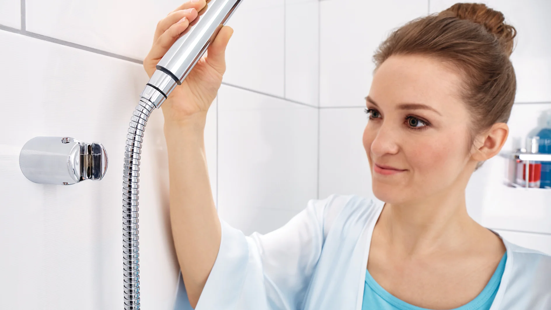 tesa-Classic-black-Spaa-40343_mood Eine Frau in einem hellblauen Top und Bademantel hält einen abnehmbaren Duschkopf in einem Badezimmer mit weiß gefliesten Wänden. Sie betrachtet den Duschkopf und bereitet sich darauf vor, ihn zu benutzen. (This text has been generated by AI)