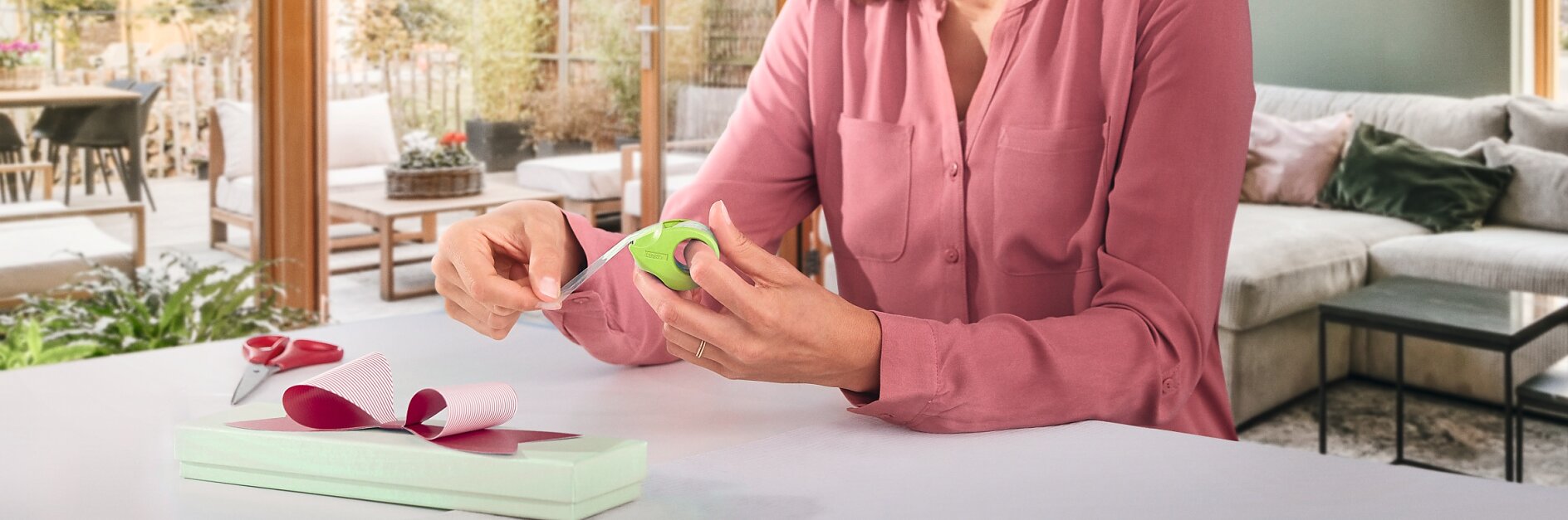 Eine Frau mit schulterlangem braunem Haar und einer rosa Bluse sitzt an einem weißen Tisch in einem modernen Wohnzimmer und verpackt eine hellgrüne Geschenkbox mit Tesa-Klebeband und verschließt sie mit einer rosa Schleife. Große Fenster geben den Blick auf einen Garten frei. (This text has been generated by AI)