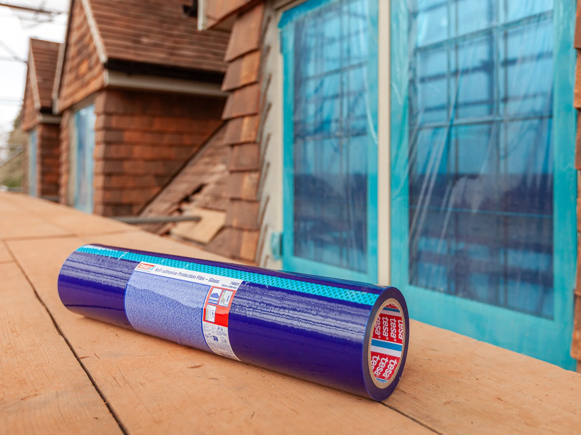 A roll of blue tesa tape adhesive membrane sits on a wooden surface outside a building under construction, with blue-framed windows and brown siding visible in the background. (This text has been generated by AI)