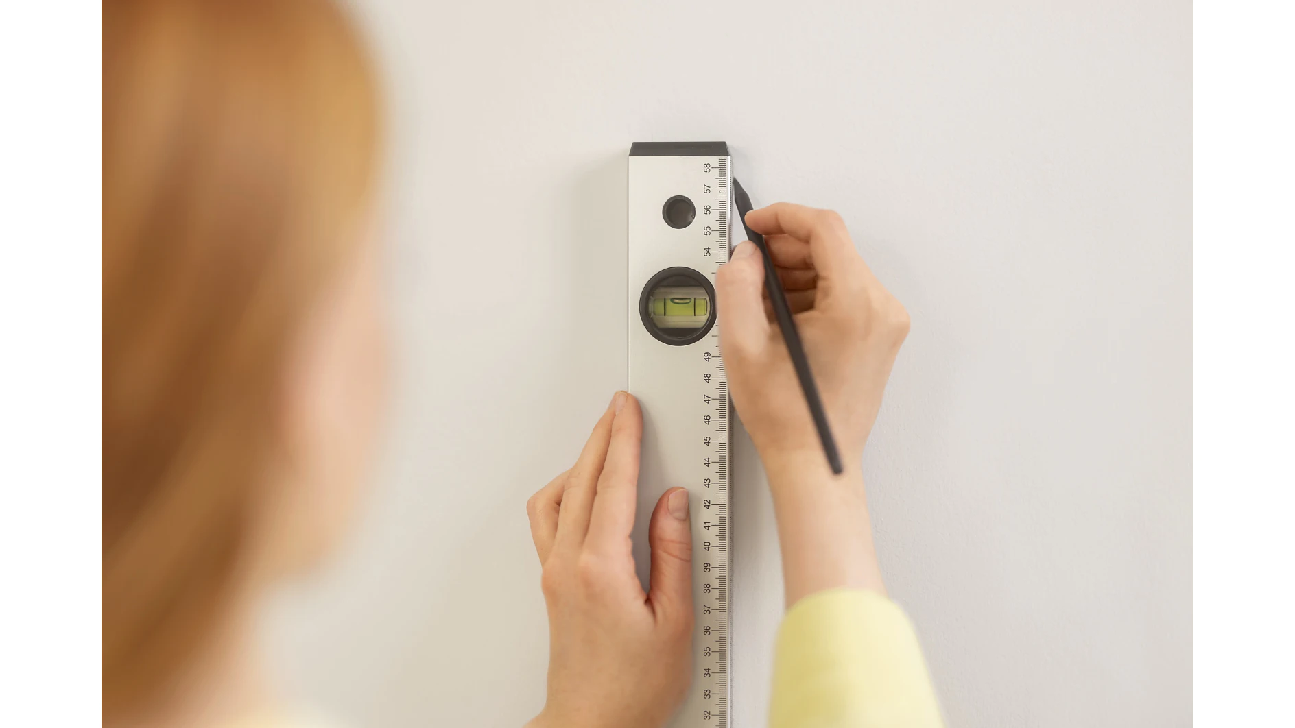 Eine Person benutzt ein Lineal mit eingebauter Wasserwaage und Tesaband, um mit einem Bleistift eine vertikale Linie auf eine weiße Wand zu zeichnen. (This text has been generated by AI)