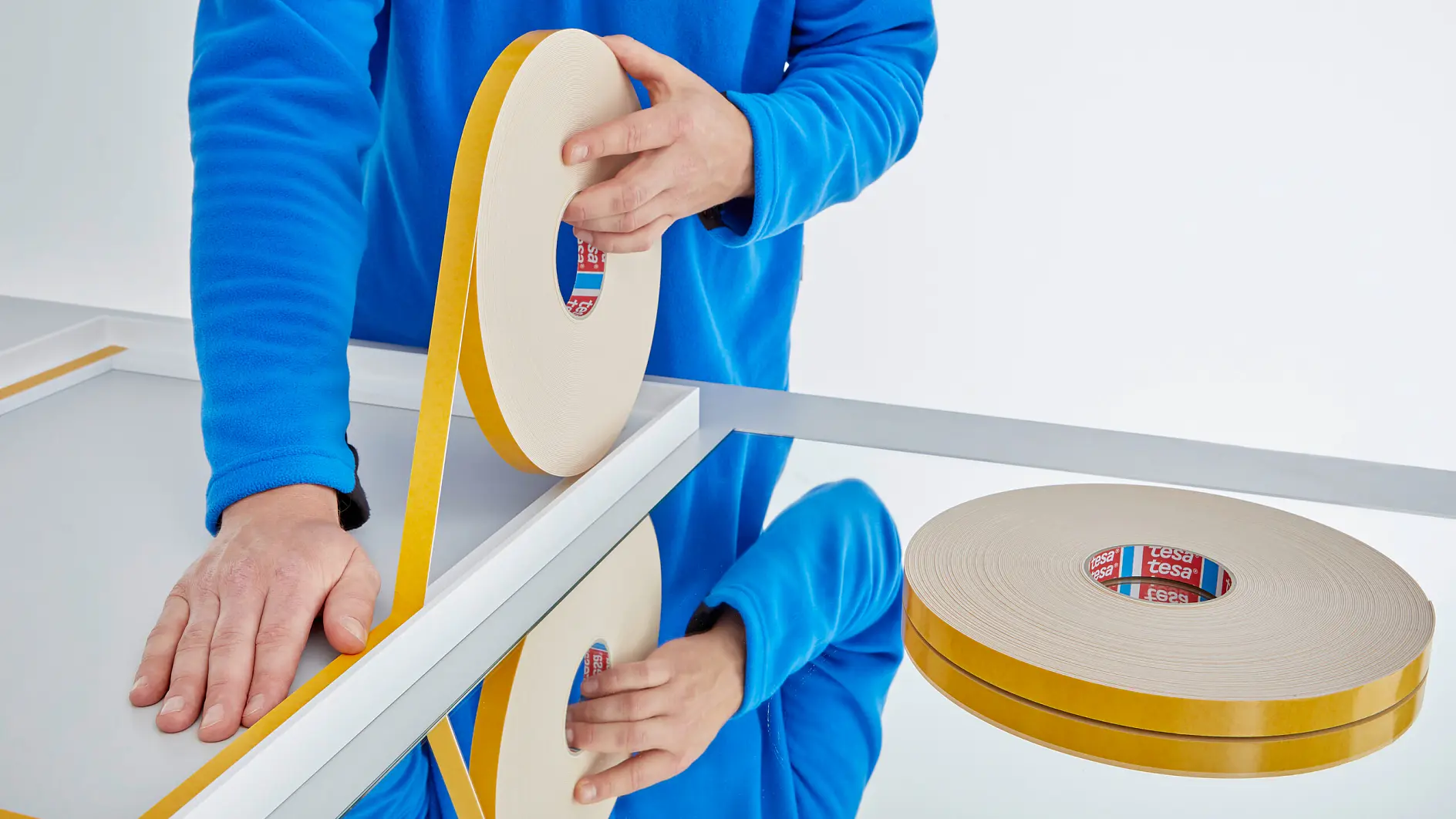 Person in blauer Jacke bringt tesa doppelseitiges Klebeband auf weißen Rahmen auf reflektierender Oberfläche in heller Innenraumumgebung an (This text has been generated by AI)