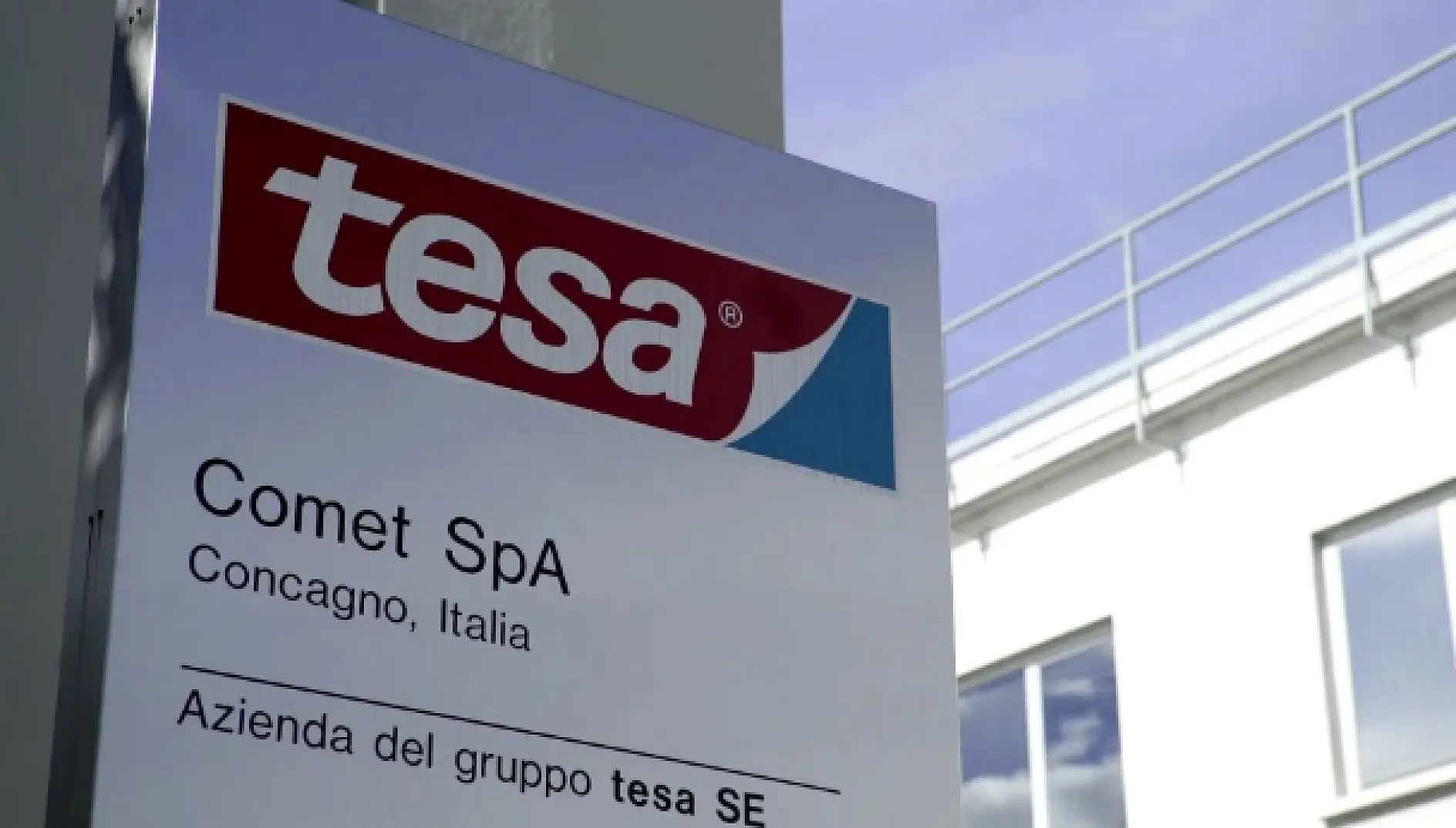 Auf einem Schild mit dem tesa-Bandlogo oben steht "Comet SpA, Concagno, Italia" und "Azienda del gruppo tesa SE". Im Hintergrund sind ein modernes Gebäude und der blaue Himmel zu sehen. (This text has been generated by AI)