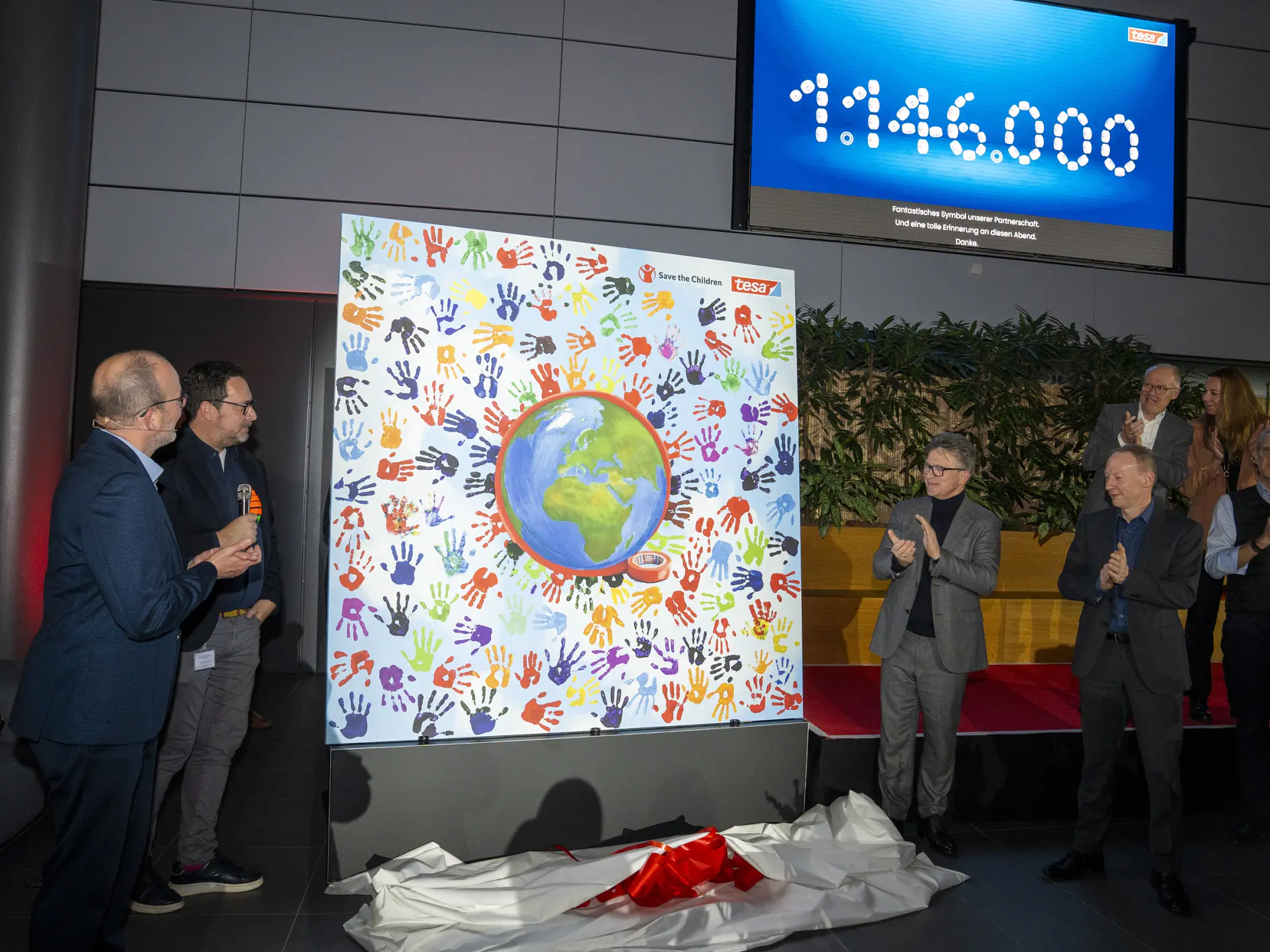 Eine Gruppe steht vor einem bunten Display mit Handabdrücken der Erde. Einige klatschen. Ein Bildschirm zeigt 1.146.000. Auf dem Boden liegen Tesafilm und ein rot-weißes Tuch. (This text has been generated by AI)