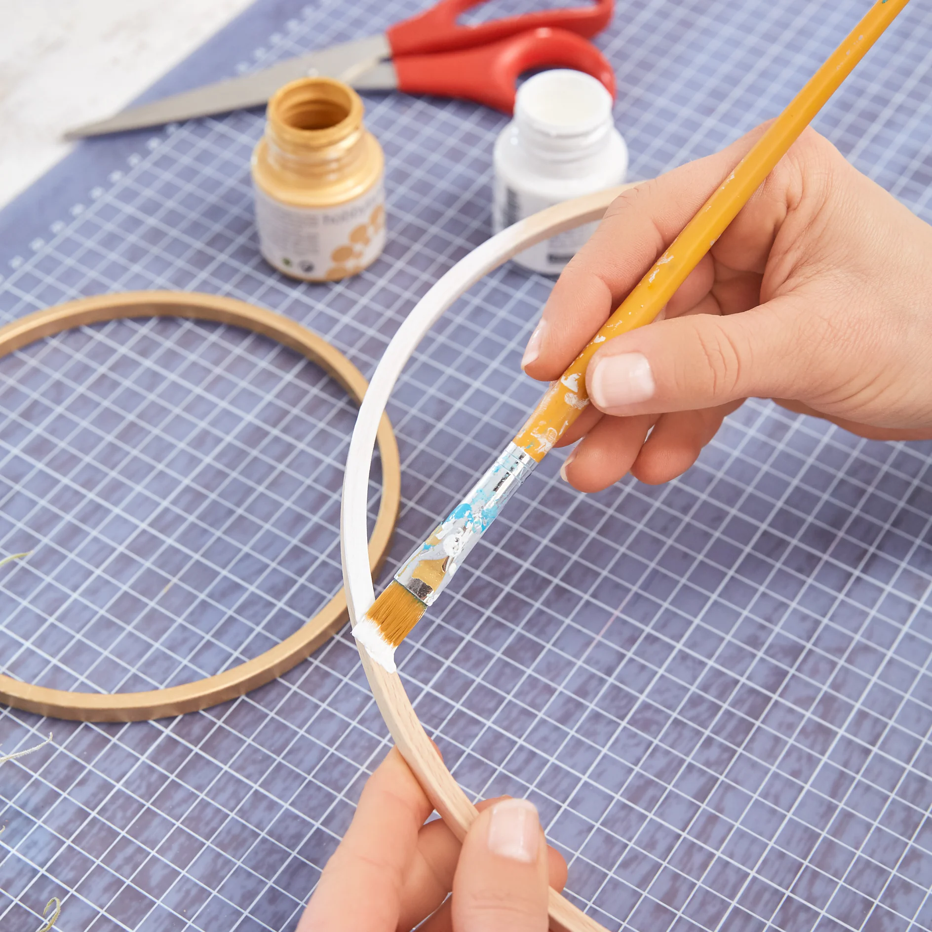 Hands painting a wooden embroidery hoop with a paintbrush. Two paint jars, one gold and one white, and red scissors are on a grid-patterned cutting mat in the background. Rolls of tesa tape can also be seen neatly placed nearby. (This text has been generated by AI)