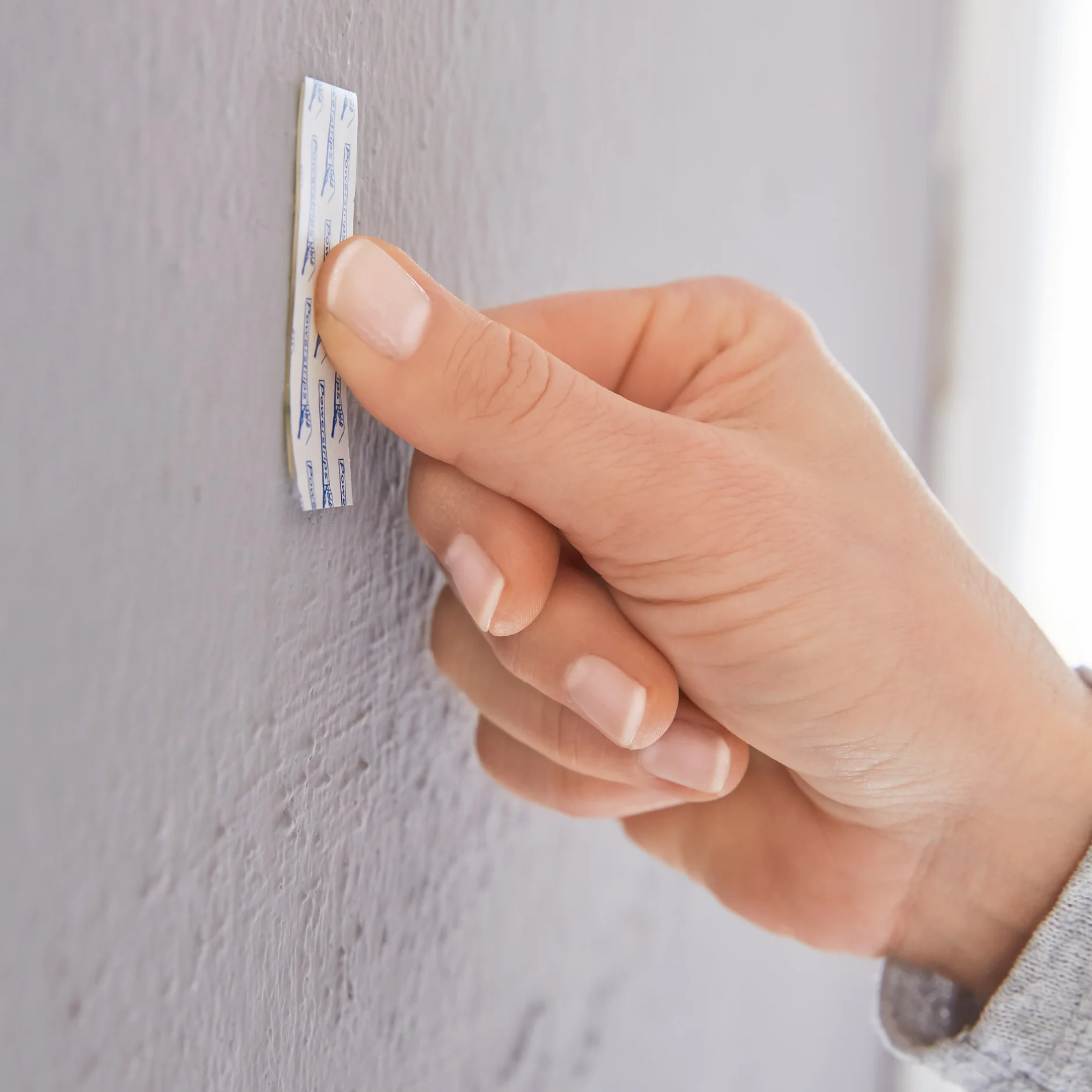 A persons hand is sticking a small tesa tape strip onto a textured gray wall. The tesa tape strip has a blue chevron pattern and a white backing. The persons thumb presses firmly on the tape to adhere it to the wall surface. (This text has been generated by AI)